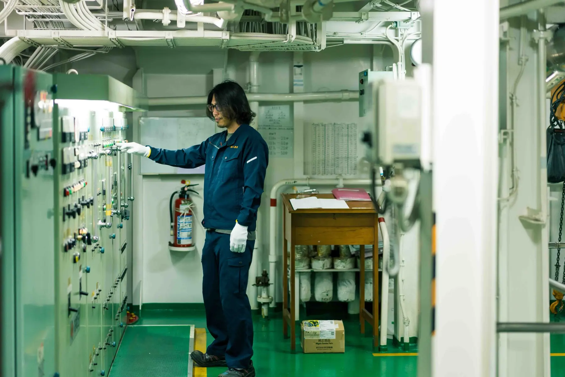 船の機関室で計器盤に向かう作業服の人